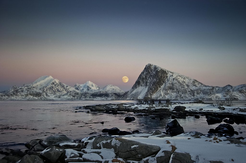 Beautiful twilight view of snow-capped mountains and full moon reflecting on a tranquil lake.